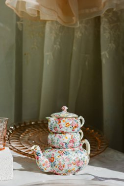 Bir çaydanlık ve fincanlar masaya istiflenir ve pencereden gelen yumuşak güneş ışığı sıcak bir ortam oluşturur..