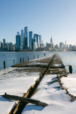 Karla kaplı bir rıhtım, kışın açık bir gökyüzünün altında görünen bir şehir silueti olan bir nehre doğru uzanıyor..