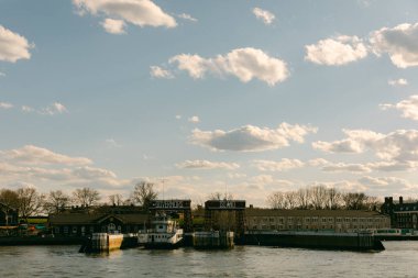 Rıhtıma bir tekne yanaştı. Binalar bulutlarla dolu mavi bir gökyüzünün altındaki nehir kıyısında. Öğleden sonra oldu. Bölgede insanlar dolanıyor..