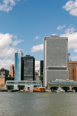 Nehir boyunca rıhtıma yakın tekneleri olan birkaç yüksek bina var. Gökyüzü parçalı bulutlu ve güneş suyun üzerinde parlıyor. Bu sahne şehirdeki kentsel yaşamı yakalıyor..