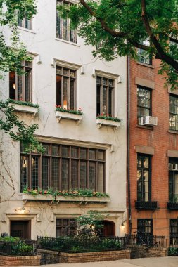 Caddede birkaç penceresi olan yüksek bir bina duruyor. Çiçekler pencere girişlerini dekore eder. Yakınlarda ikinci bir bina var. Ağaçlar bölgeye gölge düşürür.