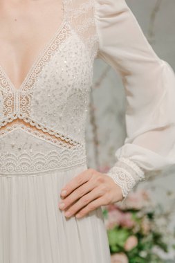 Bir gelin, güzel bir ortamda ayrıntılı bir gelinlik giyerek durur. Arka plandaki çiçeklere bakarken elini beline koyar. Sahne düğün neşesiyle dolu..