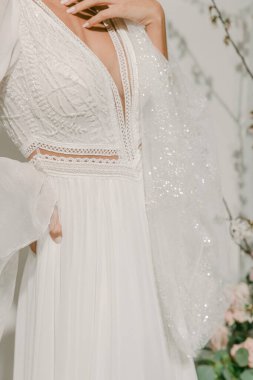 Çiçek desenli beyaz bir elbise giyen bir gelin. Elbisenin uzun kolları ve kalın bir yakası var. Arka planda çiçekler var..