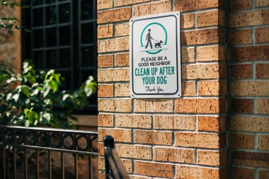 Tuğla duvara köpek sahiplerine mahalledeki evcil hayvanlarının arkasını temizlemeleri gerektiğini hatırlatan bir tabela asılıyor. Sahne, yakınlarındaki yeşilliklerle güneşli bir günü gösteriyor..