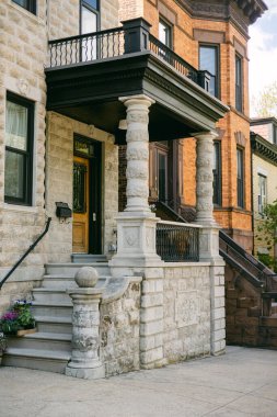 Brownstone binası ayrıntılı taş işçiliğini ve önemli bir verandayı gösteriyor. Çiçekler basamaklara oturur. Burası açık mavi gökyüzünün altındaki bir şehir..
