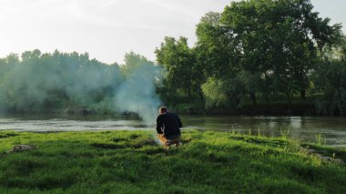 Nehir kıyısındaki bir yangının üzerine eğilmiş bir balıkçı balık çorbası hazırlıyor..
