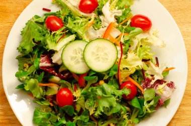 Salata yaprakları, salatalık dilimleri ve ahşap arka planda beyaz bir tabakta kiraz domatesleri.