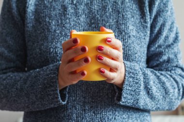 Gri desenli kazaklı kadın elinde sarı bir fincan çay tutuyor.