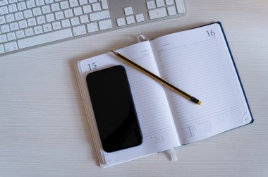 Bir akıllı telefon ve bir kalem açık bir günlüğün üzerinde boş kağıt sayfalarıyla birlikte duruyor, günlüğün yanında bir klavye var..