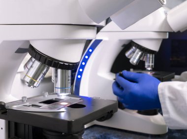 Laboratuvarda iki büyük mikroskop mikroskop, mikroskop altında numuneler, arka planda mavi eldivenli bir adam mikroskobun arkasında çalışıyor..