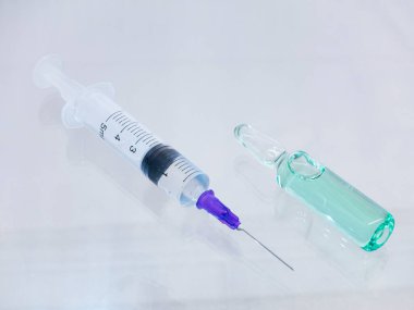 İçinde açık bir iğne ve temiz bir sıvı bulunan bir şırınga yanında yeşil bir sıvı bulunan bir ampul yatar..