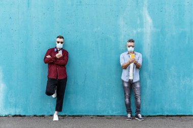 Havalı adamlar sosyal mesafeyi koronavirüs salgını sırasında cep telefonlarını kullanırken ve yüz maskesi takarken, arkadaşlar video izliyor ve akıllı telefonla mesajlaşıyorlar.