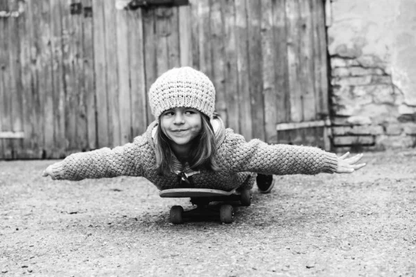 Adorable little girl in winter wool clothes lying skateboard flies like a plane outdoors in backyard - Smiling little girl playing with skateboard - Vintage black and white version