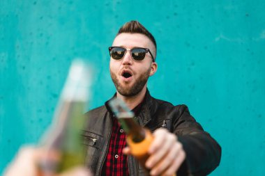 Sakallı, havalı bir adam, en iyi arkadaşıyla dışarıda bira içiyor, arka planda mavi bir duvar var. Deri ceket giyen, bira şişeleriyle kadeh kaldıran hippi adamın elleri. Dostluk kavramı, erkek yüzüne odaklan.