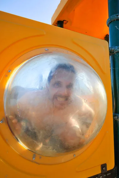 A man makes funny faces behind the clear plastic porthole in the kids\' gym on the beach during summer vacation - Guy enjoying life on the beach making comic faces - Forever young concept