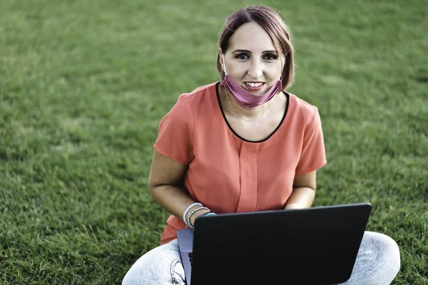 Woman 40 years old working outdoors with laptop during coronavirus outbreak - Smiling female entrepreneur sitting on lawn with personal computer while looking camera - Remote work concept into nature