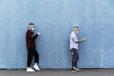 Sosyal mesafeyi korumak ve yüz maskesi takmak için cep telefonu kullanan havalı adamlar, Coronavirus salgını sırasında tek sıra halinde video izleyip akıllı telefonla mesajlaşan arkadaşlar.