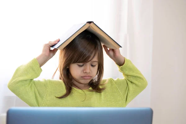 Sad elementary schooler girl studying at home with a book on her head because she is tired and angry - Little girl studying online and using internet lessons and school book - Digital divide concept