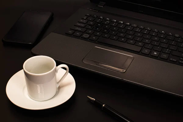 A cup of coffee, a computer, a pen and a phone on a black background. Workplace of a businessman