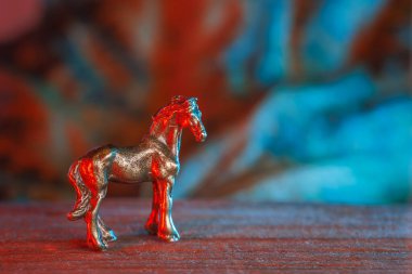 Masanın üstünde minyatür metalik at, kırmızı ve mavi sinematik ışıkla yıkanmış, bokeh arkaplanlı sığ odak, parlak desenli yüzey, nostaljik