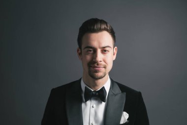 Smokinli genç beyaz bir adamın stüdyo resmi, kameraya bakıyor, stüdyo arka planına bakıyor.