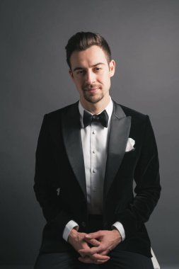 Smokinli genç beyaz bir adamın stüdyo resmi, kameraya bakıyor, stüdyo arka planına bakıyor.