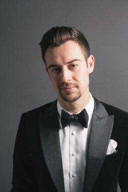 Smokinli genç beyaz bir adamın stüdyo resmi, kameraya bakıyor, stüdyo arka planına bakıyor.