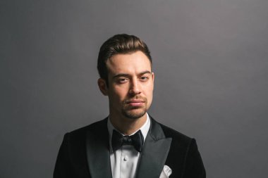 Smokinli genç beyaz bir adamın stüdyo resmi, kameraya bakıyor, stüdyo arka planına bakıyor.