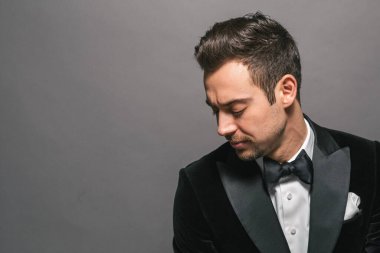 Smokinli genç beyaz bir adamın stüdyo portresi, gömleğin düğmelerini açıyor, düz stüdyo arka planına bakıyor.