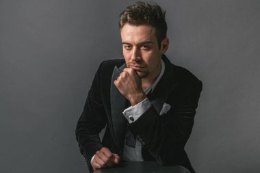 Smokinli genç beyaz bir adamın stüdyo portresi, çene üstüne el ele, ciddi ciddi kameraya bakıyor, düz stüdyo arka planına karşı