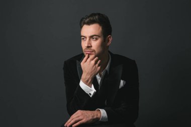 Smokinli genç beyaz bir adamın stüdyo portresi, el pençe divan duran, ciddi bir şekilde yandan bakan, düz stüdyo arka planı