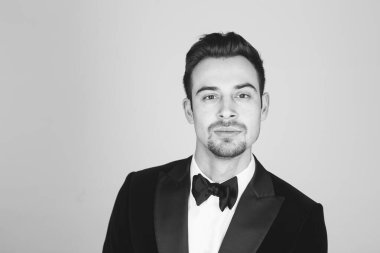 Smokinli genç beyaz bir adamın stüdyo resmi, kameraya bakıyor, stüdyo arka planına bakıyor.