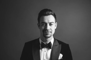 Smokinli genç beyaz bir adamın stüdyo resmi, kameraya bakıyor, stüdyo arka planına bakıyor.