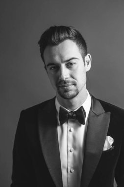 Smokinli genç beyaz bir adamın stüdyo resmi, kameraya bakıyor, stüdyo arka planına bakıyor.