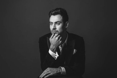 Smokinli genç beyaz bir adamın stüdyo portresi, el pençe divan duran, ciddi bir şekilde yandan bakan, düz stüdyo arka planı