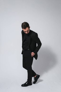 Siyah ceketli genç beyaz bir adamın stüdyo portresi, el ele bakıyor, düz stüdyo arka planında duruyor.