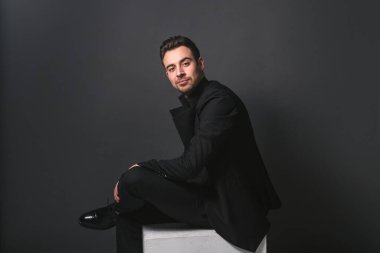 Siyah ceketli beyaz bir adamın stüdyo portresi, kameraya bakıyor, stüdyo arka planında oturuyor.