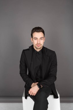 Siyah ceketli beyaz bir adamın stüdyo portresi. El ele tutuşmuş, kameraya bakıyor, stüdyo arka planında oturuyor.