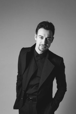 Siyah ceketli genç beyaz bir adamın stüdyo portresi, kameraya bakıyor, elleri cepte, düz stüdyo arka planında duruyor.