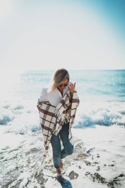 Omuzlarında battaniyeyle deniz kıyısında çıplak ayakla yürüyen zarif sarışın bir kadının tam boy portresi. Şık güneş gözlüklü hippi kız deniz kenarında tatilinin tadını çıkarıyor..