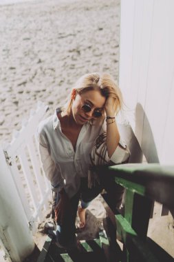 Moda güneş gözlüklü sarışın bir kadının, omuzlarında ekoseli bir kıyafetle plaj evinin merdiveninin dibinde kameraya bakarken çekilmiş en güzel fotoğrafına bakın.. 