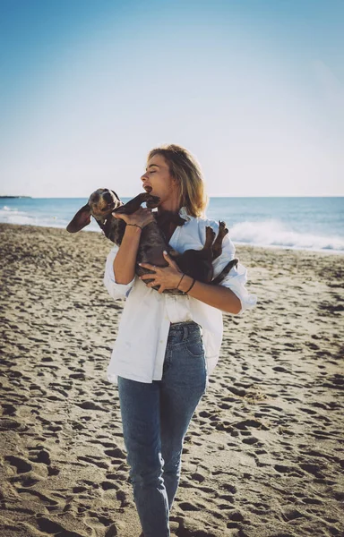 海の海岸の背景にビーチに立っている間 陽気なブロンドの女性は彼女の素敵なペットの犬を抱きかかえています 白いシャツの魅力的な白人女性と青いジーンズ楽しいですザ日オフとともに彼女の犬 — ストック写真