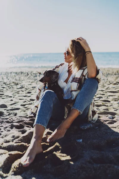 美しいモデルは 彼女の美しい犬と座っている間にブロンドの女性が笑顔に見え ビーチサイドでリラックスします 陽気な女性座って素足で砂の上に彼女の子犬楽しんで晴れた日にザ海辺で — ストック写真