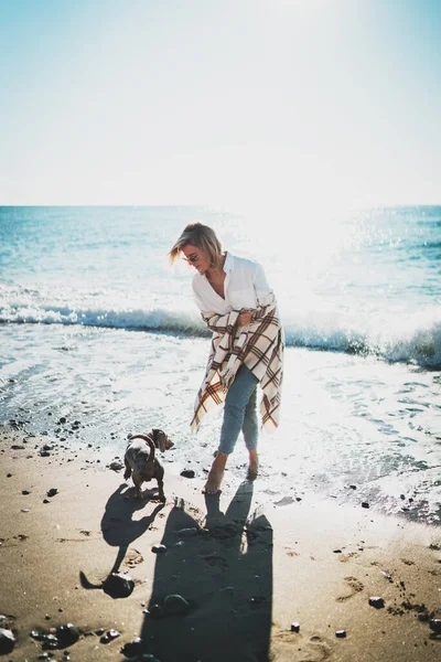 海岸で犬と遊んでいる魅力的なブロンドの女性 晴れた日に彼女のペット犬と一緒に歩いて彼女の肩の上に再生されたカジュアルな服の裸足の女性 週末はビーチで一杯 — ストック写真