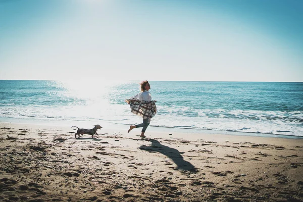 晴れた日にペットの犬と一緒に海沿いを走っている幸せなブロンドの女性 陽気な白人女性で青ジーンズともに彼女の肩遊びとともに彼女の犬でザビーチサイド — ストック写真