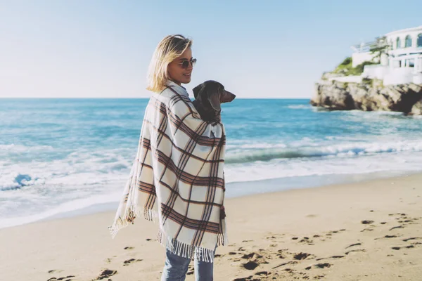青いジーンズを着た魅力的なブロンドの女性は 海岸のぼやけた背景に彼女の腕の中で彼女の犬と一緒に立っている彼女の肩の上に再生されます 学生女の子歩くザビーチサイドとともに彼女のパップ — ストック写真