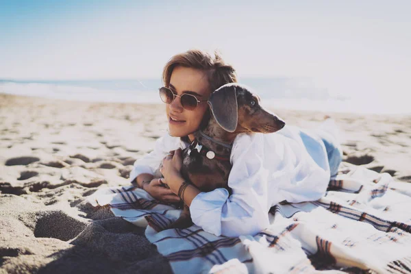 暖かい晴れた日に砂浜の毛布の上に横たわっている間 彼女のかわいい犬を愛して抱きしめるカジュアルな服の魅力的なブロンドの女性 ヒップスター女の子過ごすスペア時間とともに彼女のペット犬オンザビーチ — ストック写真
