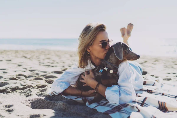 Cheerful female in casual outfit laughing and kissing dog while lying on a sand beach at the day off. Young woman is looking at her cute puppy while relaxing on a warm sunny day outdoors.