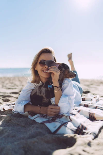 Веселая Женщина Повседневной Одежде Смеется Обнимает Собаку Лежа Песчаном Пляже — стоковое фото