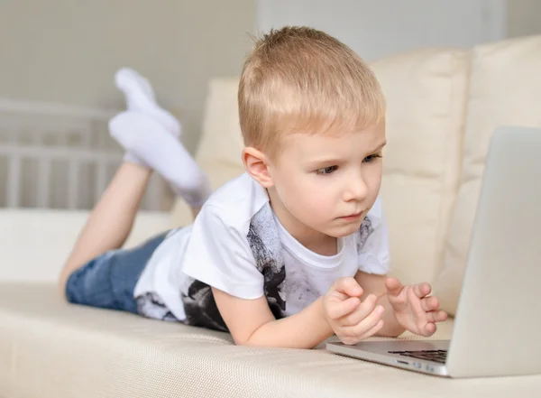 Retrato de un niño pequeño estirado en el sofá delante de la computadora portátil —  Fotos de Stock